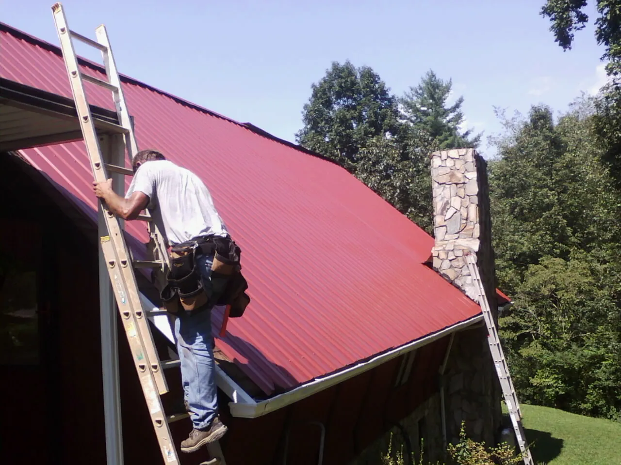 Roofer on a ladder beside a red metal roof and stone chimney — Soffit & Fascia Repair by Roofing Brothers in Huntly